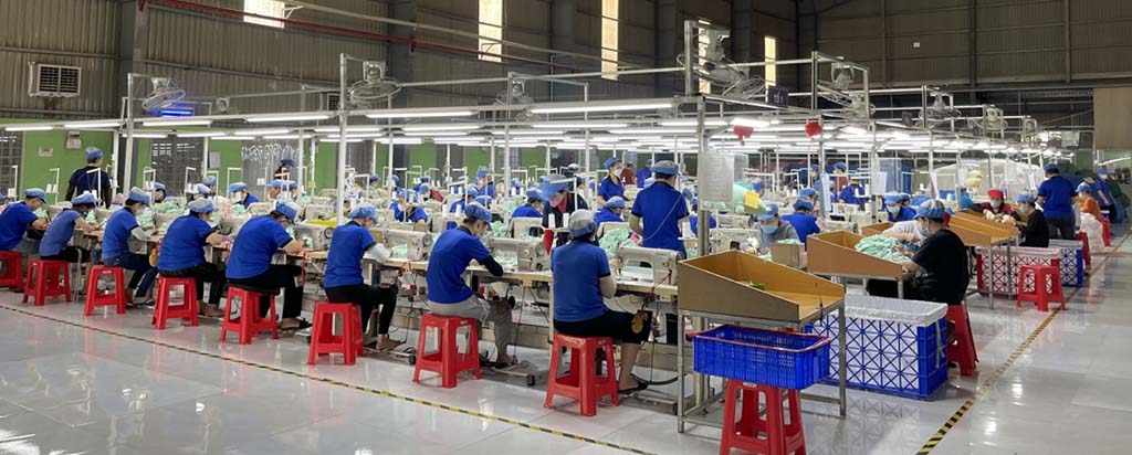 Workers in blue uniforms and caps assembling plush toys on sewing lines inside a large, well-lit toy manufacturing factory.