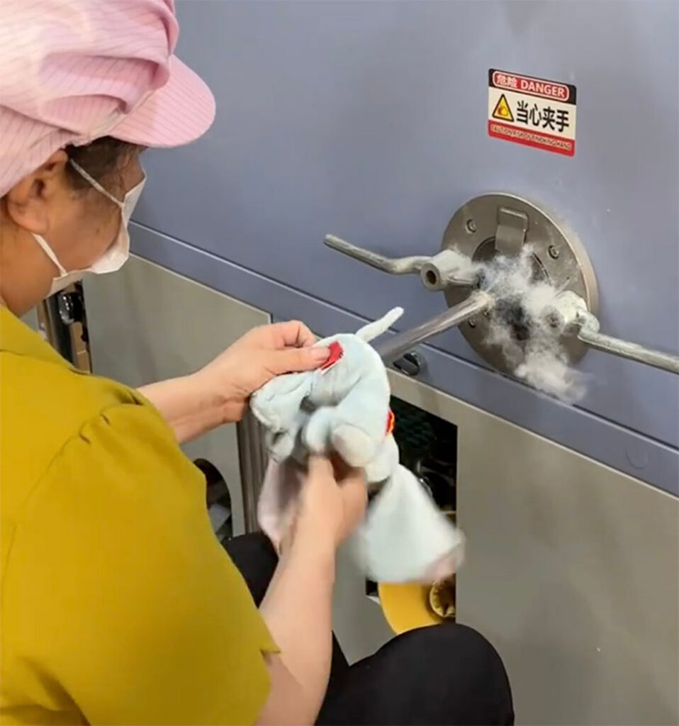 Worker using an industrial stuffing machine to fill a plush toy with cotton material during the manufacturing process.