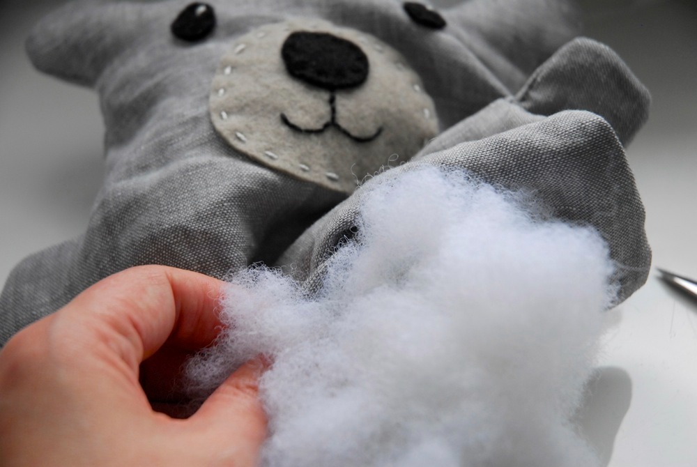 Hand stuffing a grey bear plush toy with soft white polyester fiberfill, showing recycled stuffing material in use.