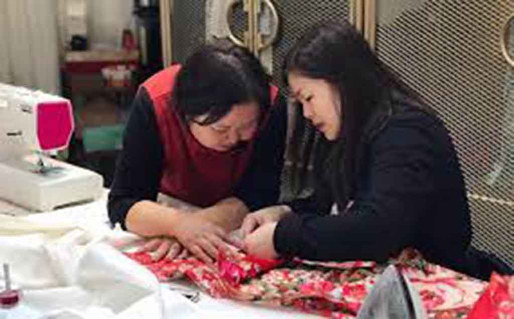 Two women carefully sewing colorful fabric together at a workshop table with a sewing machine nearby.