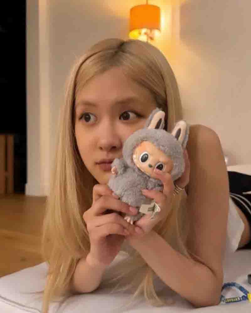 A young woman with long blonde hair holding a small Labubu plush toy in a gray bunny outfit while lying indoors.