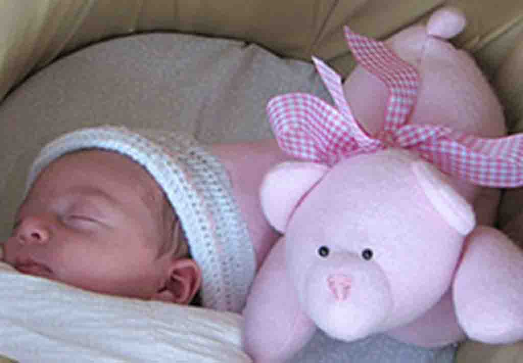 Sleeping newborn beside soft pink teddy bear with checkered ribbon bow.