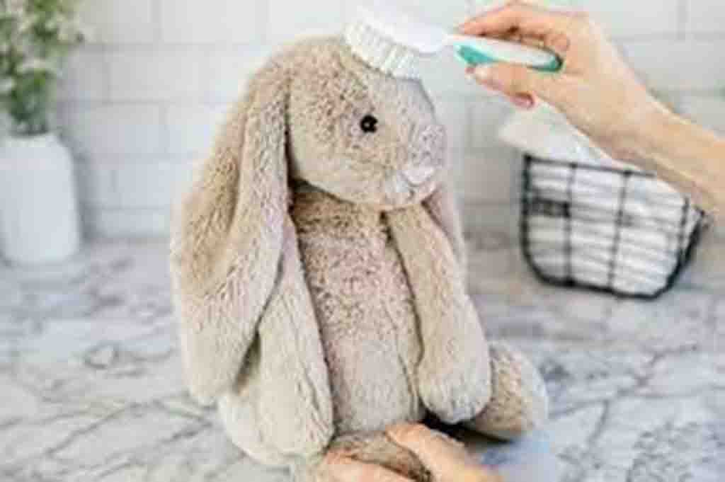 Hand using a brush to clean a light brown stuffed bunny toy on a marble surface.