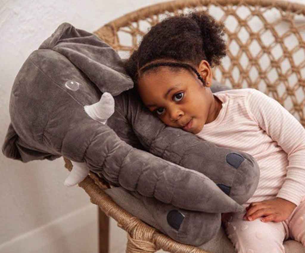 Young girl cuddling a large gray elephant plush toy while sitting in a wicker chair.