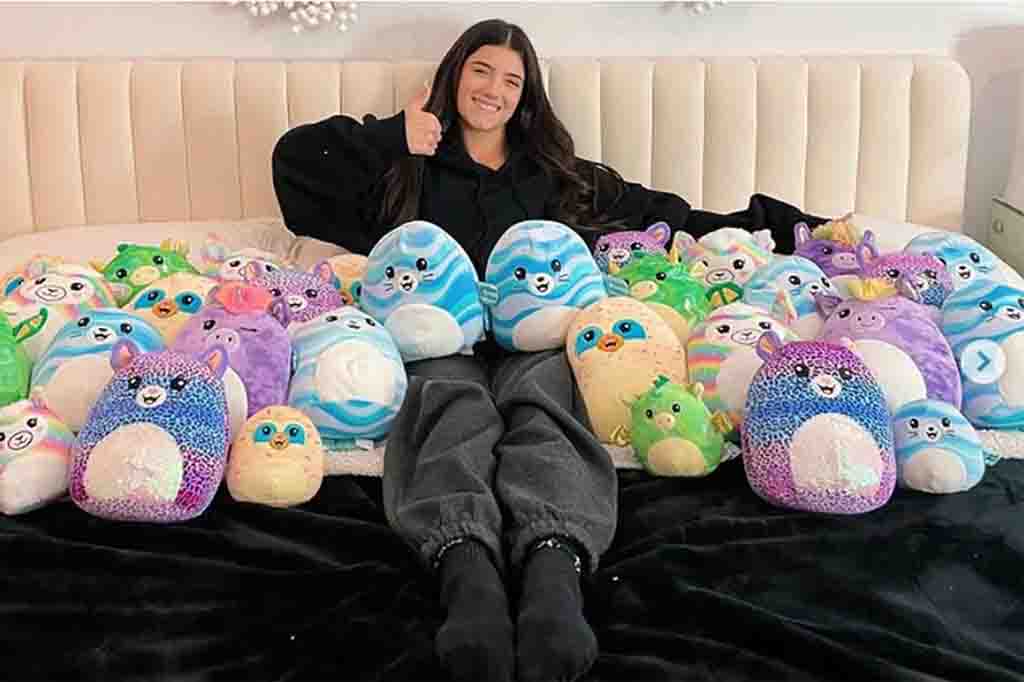 Smiling girl giving thumbs up while sitting on a bed surrounded by colorful Squishmallow plush toys.