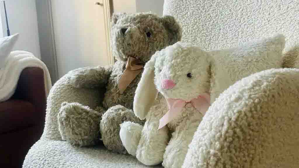 Brown teddy bear and white bunny plush with ribbons sitting on a soft chair.