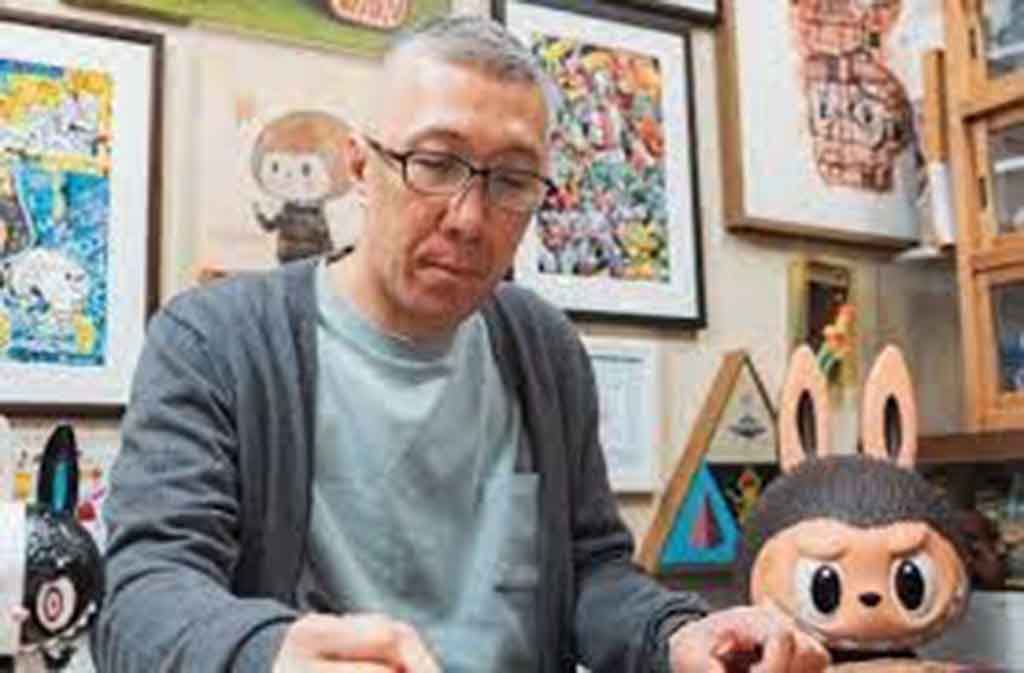 A man sketching at a desk surrounded by Labubu figures and framed artworks in a creative studio.