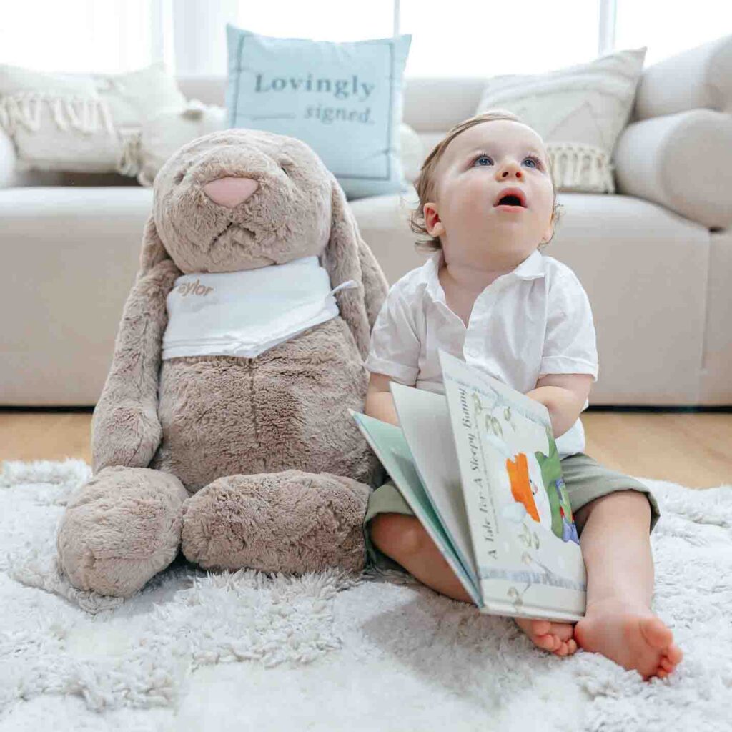 Toddler sitting on a rug next to a large plush bunny, reading a picture book, with a "Lovingly signed" pillow in the background.