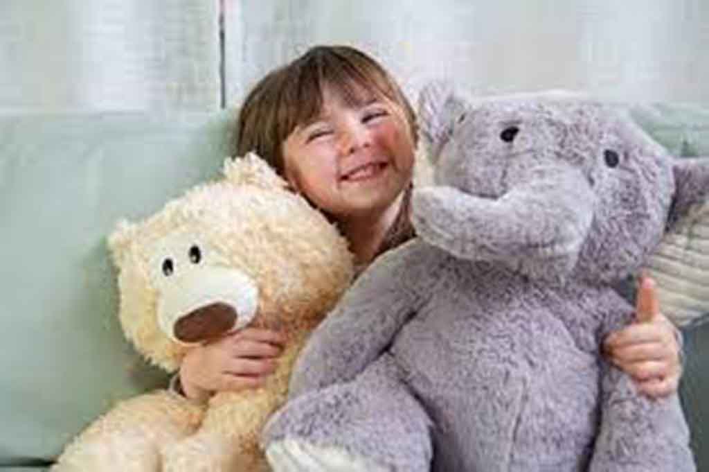 Smiling young girl sitting on a couch while hugging a teddy bear and a large elephant stuffed animal.