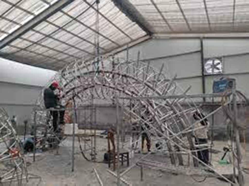 Workers assembling a large metal framework for giant stuffed animal structure inside warehouse.