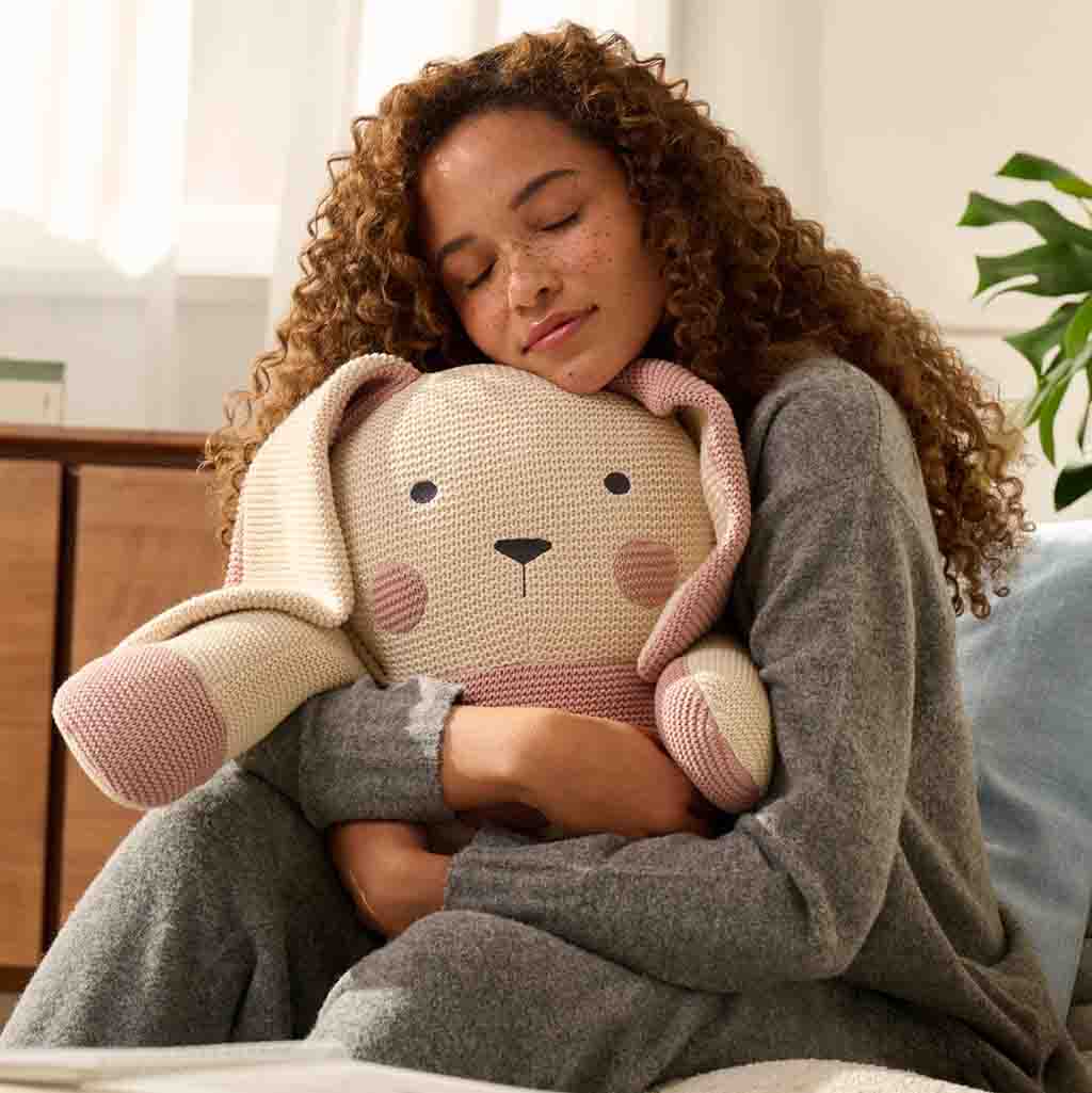 Woman hugging a soft, knitted bunny-shaped plush toy with long ears, smiling peacefully in a cozy setting.