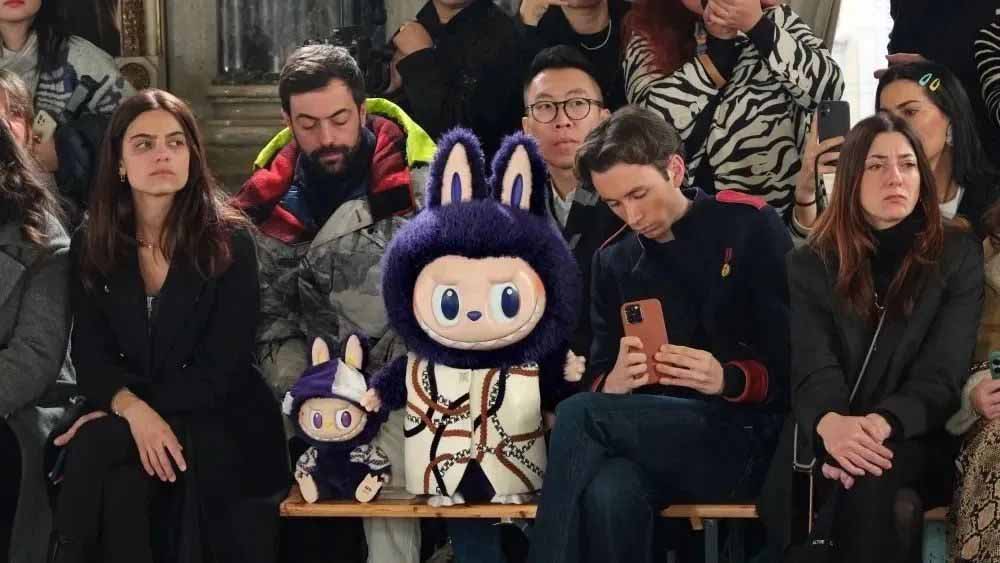 Large Labubu plush toy seated in the front row of a fashion show alongside attendees, showcasing its cultural influence.