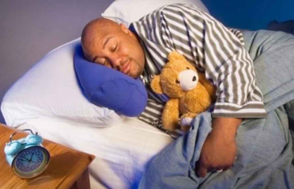 Man in striped pajamas sleeping peacefully while hugging a small teddy bear in bed.