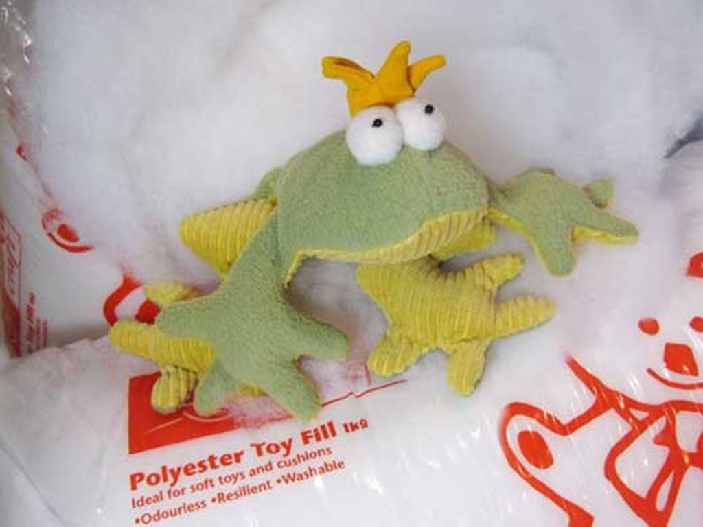 Green plush frog with a yellow crown sitting on polyester toy filling bags, used for stuffing soft toys.