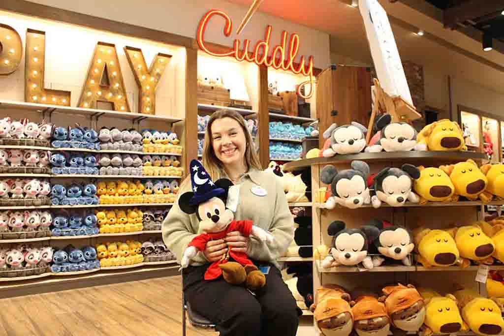 Woman holding Mickey Mouse plush in Disney store surrounded by colorful plush toys, with “Play” and “Cuddly” signs highlighting the retail display.