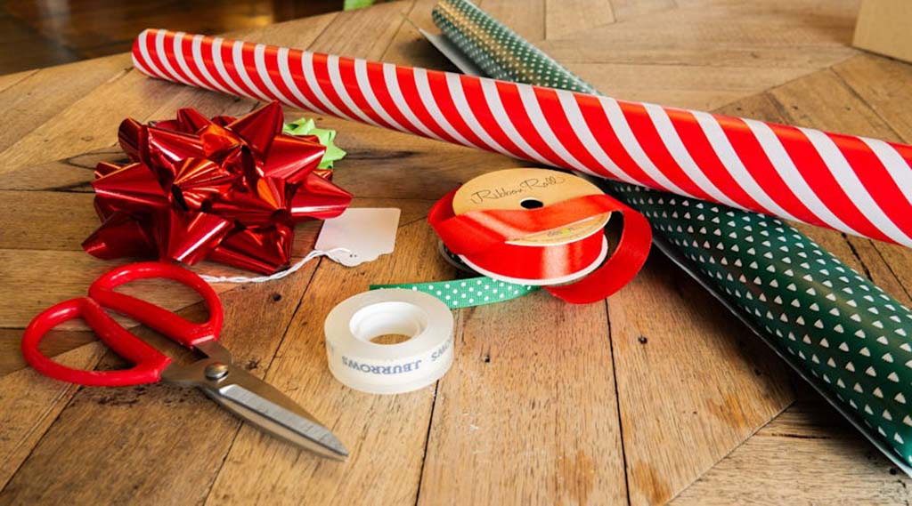 Wrapping paper rolls, ribbons, scissors, tape, and a decorative bow arranged on a wooden table for wrapping stuffed toys.