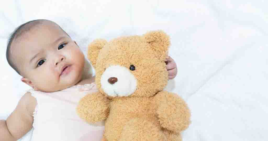 Cute baby lying on a white bed, cuddling a soft brown teddy bear while looking at the camera.