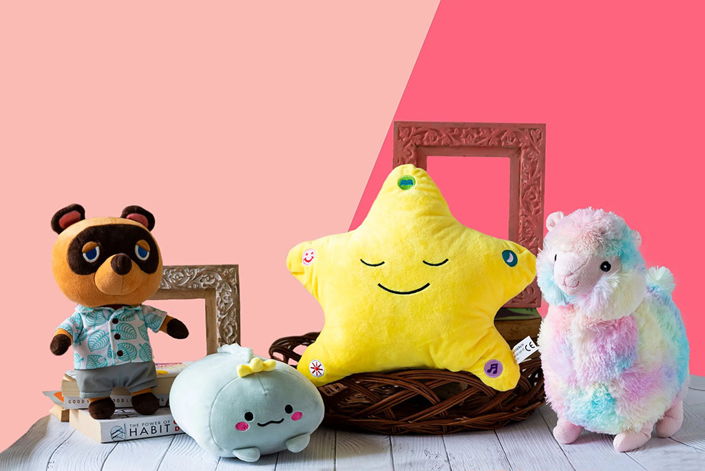 A group of colorful plush toys including a bear in a shirt, a smiling star, a rainbow llama, and a light-blue plush with a bow, displayed against a pink background.