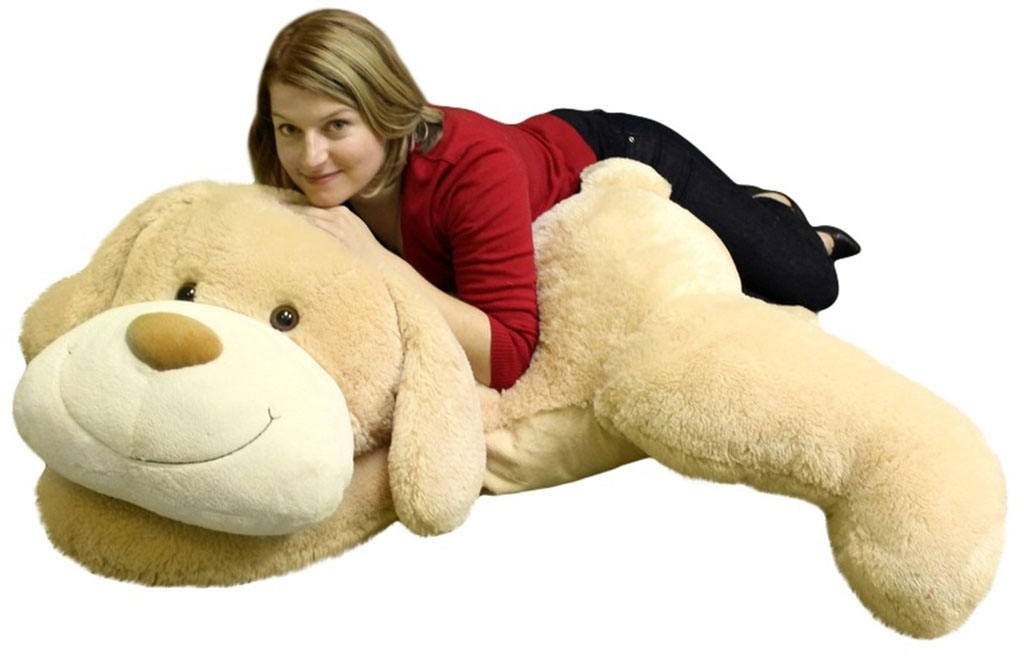 Smiling oversized beige plush dog on the floor with a woman in a red sweater lying across it.