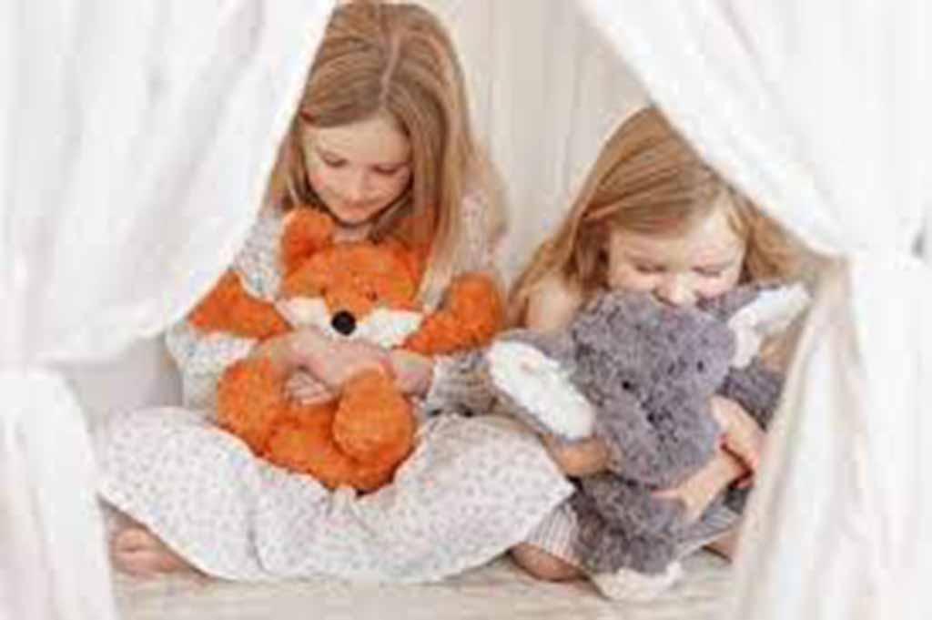 Two young girls sitting in a tent while hugging stuffed animals, one holding an orange fox plush and the other a gray elephant plush.