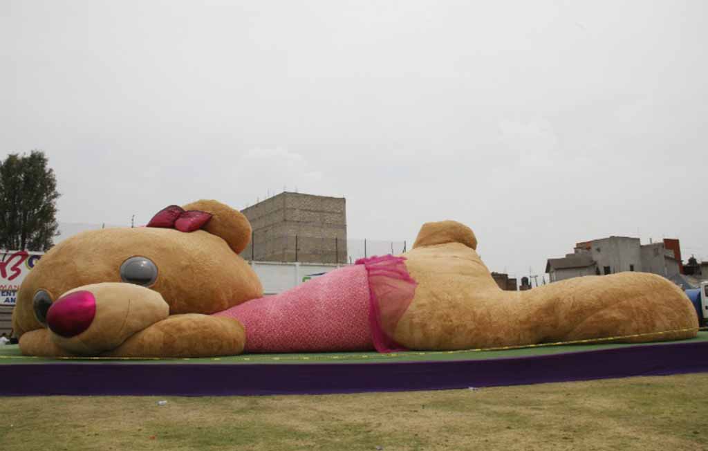 Giant teddy bear wearing a pink skirt and bow, lying on its side outdoors in a city setting.