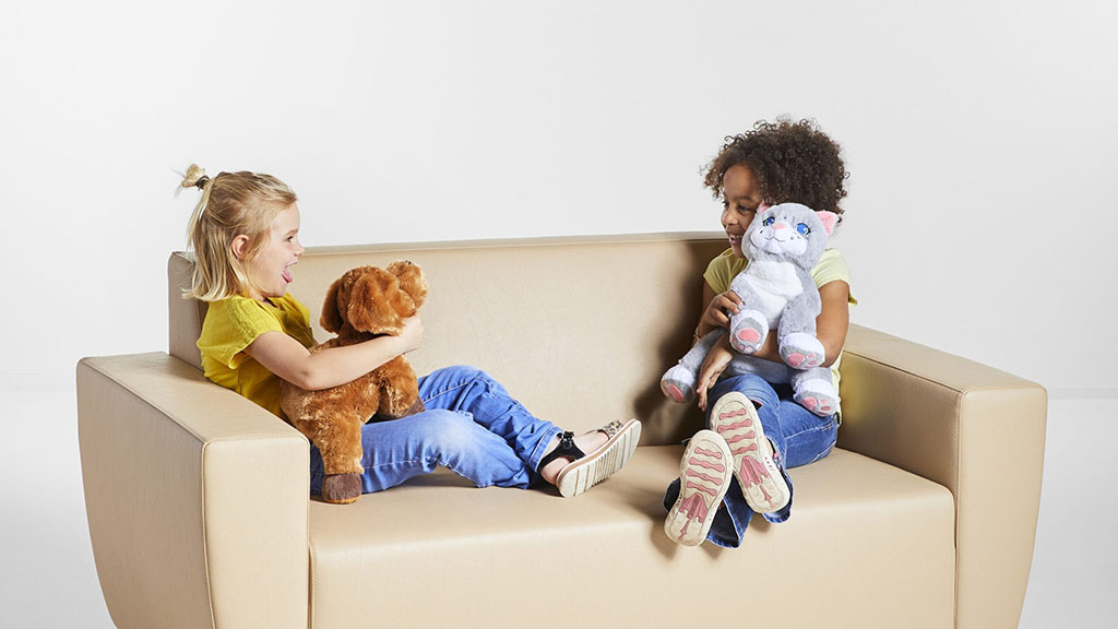 Two young children sitting on a beige couch, happily playing with stuffed animals, one holding a brown dog plush and the other a white cat plush.