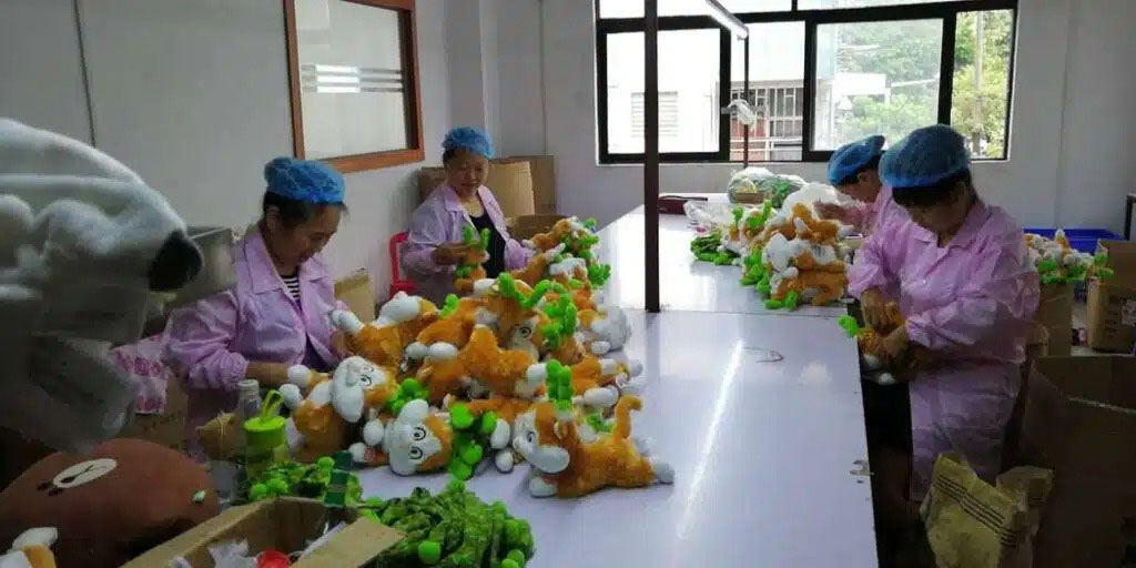 Workers in a plush toy factory sewing and assembling stuffed animals while wearing uniforms and hairnets.