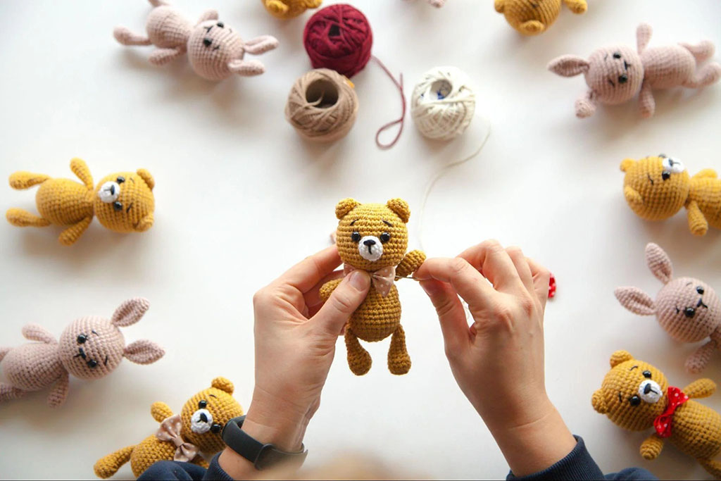Person sewing details on a small crochet bear surrounded by handmade crochet bunnies and bears with yarn balls on the table.