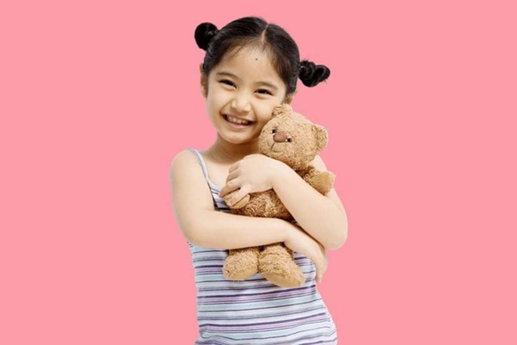 Happy young girl in striped dress hugging a small brown teddy bear against a pink background.