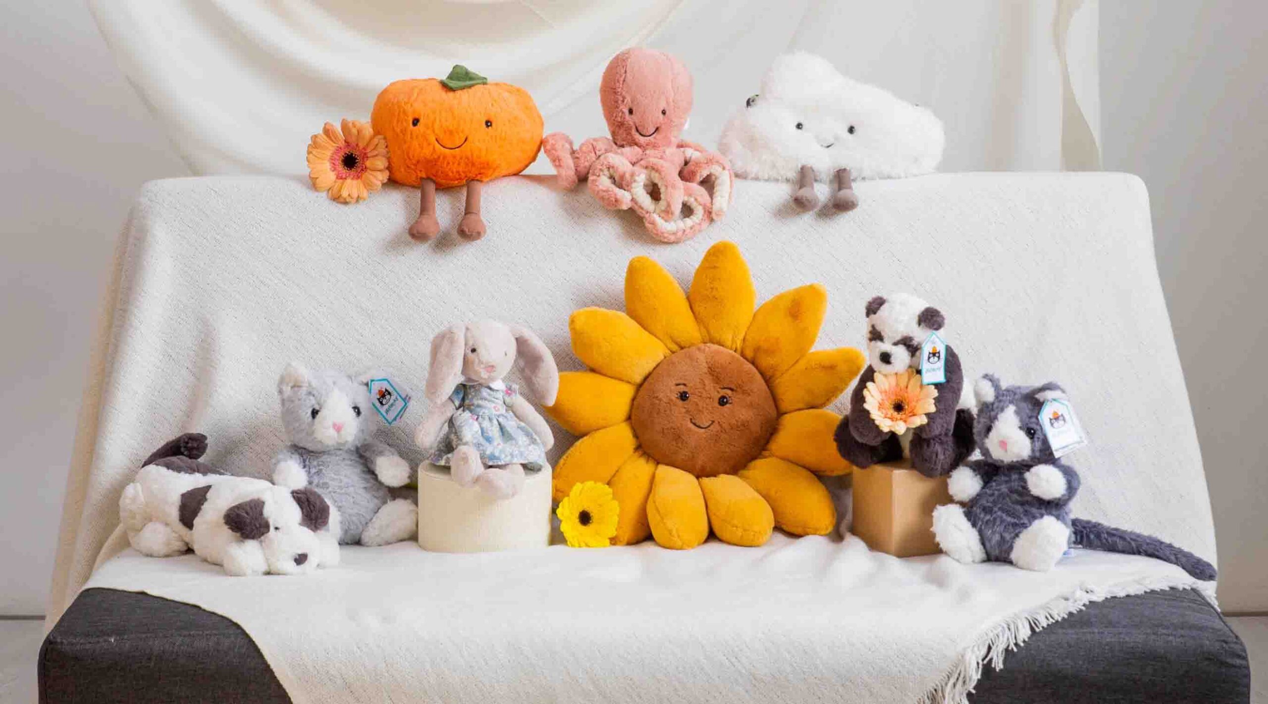 Assorted plush toys including a pumpkin, octopus, cloud, sunflower, bunny, panda, dog, and cats arranged on a white sofa.