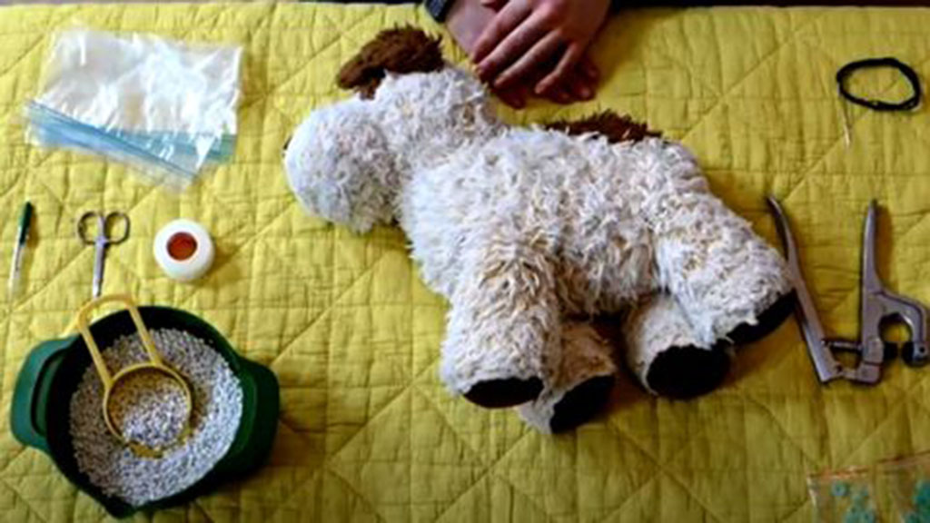 DIY setup for making a weighted stuffed animal with a plush dog, plastic pellets, scissors, tape, and sewing tools arranged on a yellow quilt.