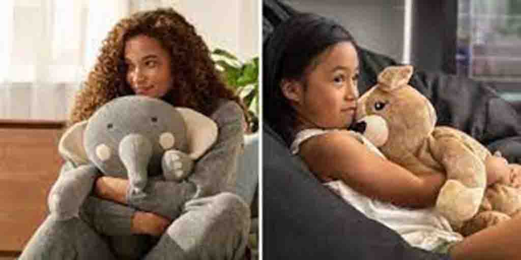 Girl cuddling gray elephant plush and another girl hugging brown kangaroo plush while sitting indoors.