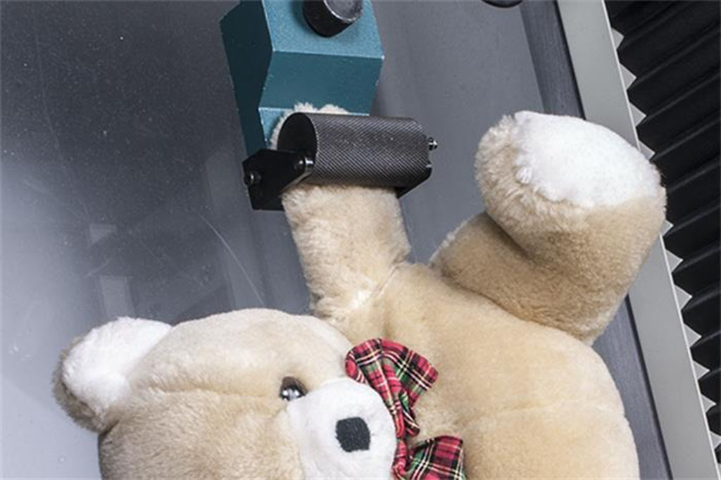 Teddy bear undergoing a mechanical safety strength test with its arm clamped in a durability testing machine.