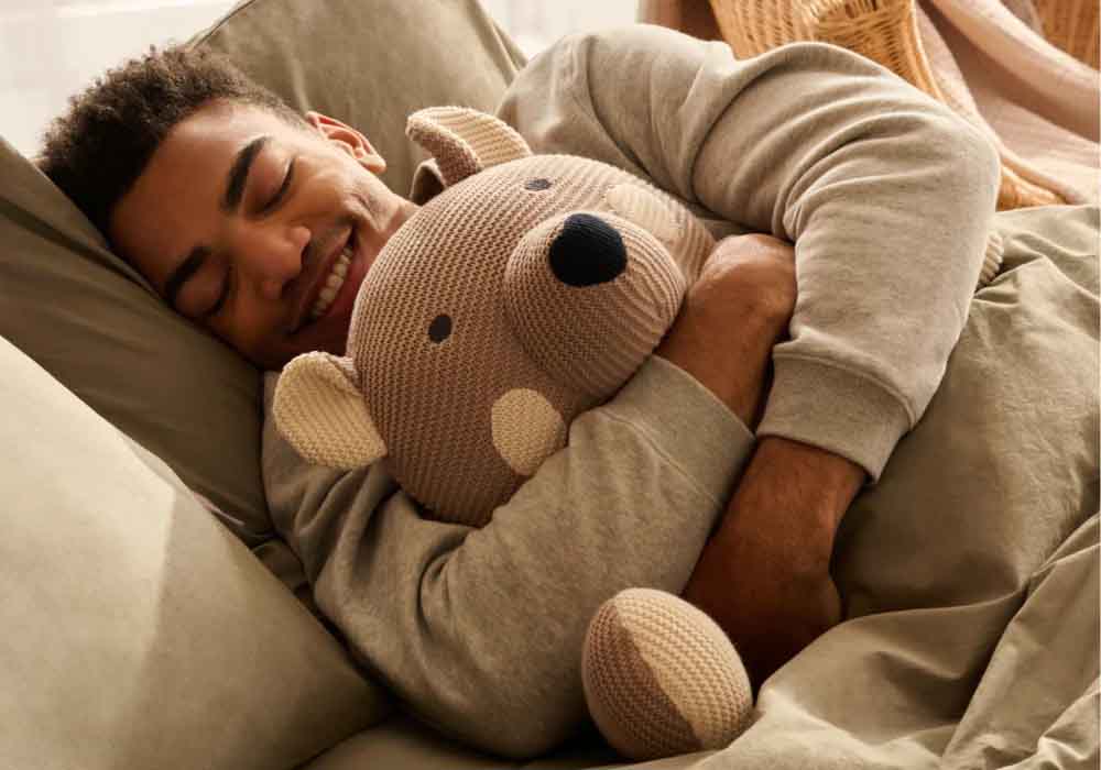 Man happily cuddling a knitted bear plush toy in bed, smiling peacefully.
