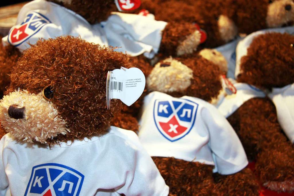 Brown teddy bears wearing white shirts printed with a blue and red sports logo, displayed in bulk for custom merchandise.