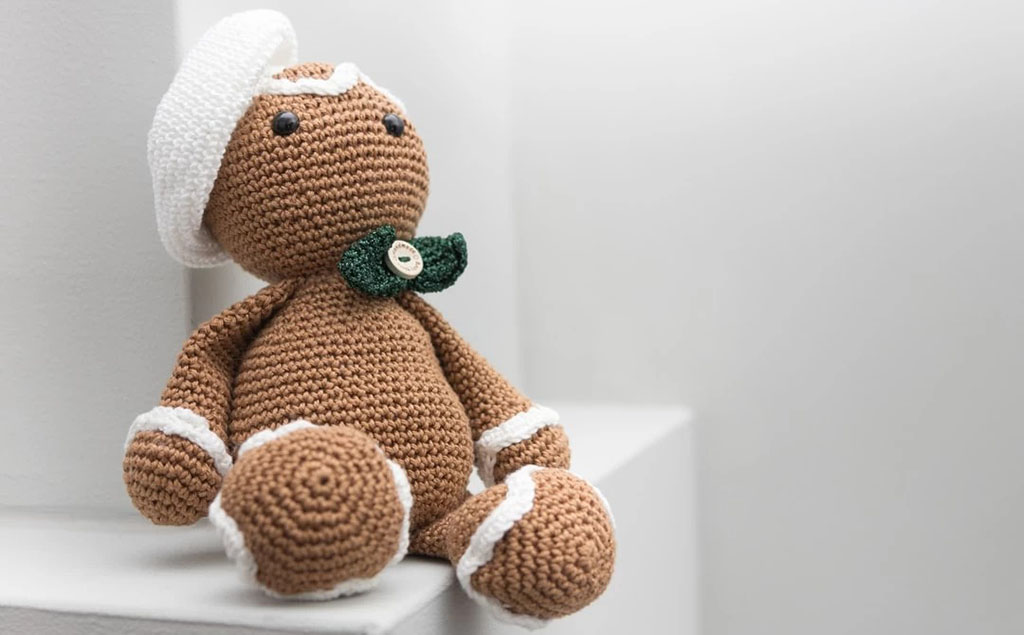 A brown crochet plush doll with a white hat, white trim accents, and a green bow tie with a button center, sitting against a minimal white background.