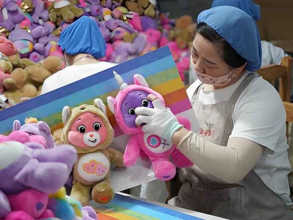 Worker in a plush toy factory inspecting colorful stuffed animals with a rainbow background for quality control.