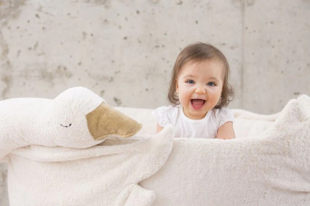 Smiling baby sitting beside a giant white goose plush toy with a beige beak, creating a cozy and playful scene.