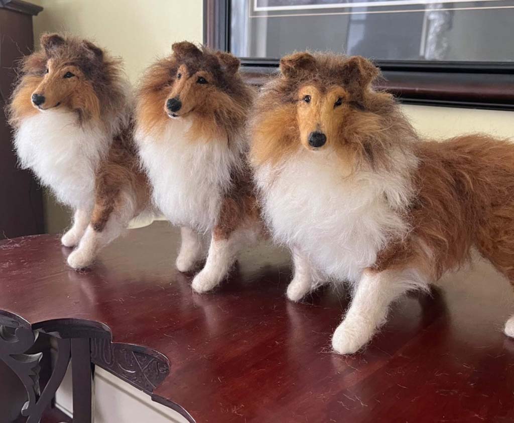 Three realistic collie stuffed animals with brown and white fur standing on a polished wooden table.