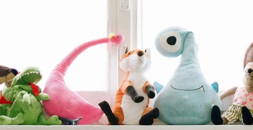 A group of plush toys including a green dinosaur, pink creature, orange fox, and a blue one-eyed monster sitting together on a windowsill.