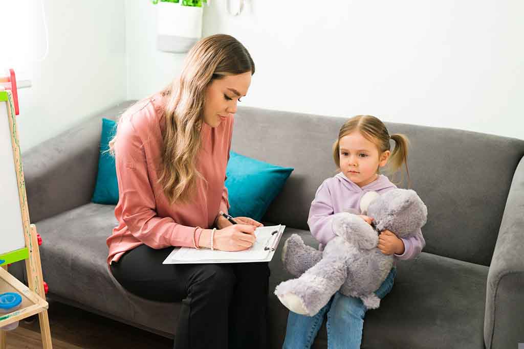 Young girl holding a gray teddy bear during a therapy session while a female therapist takes notes on a clipboard.