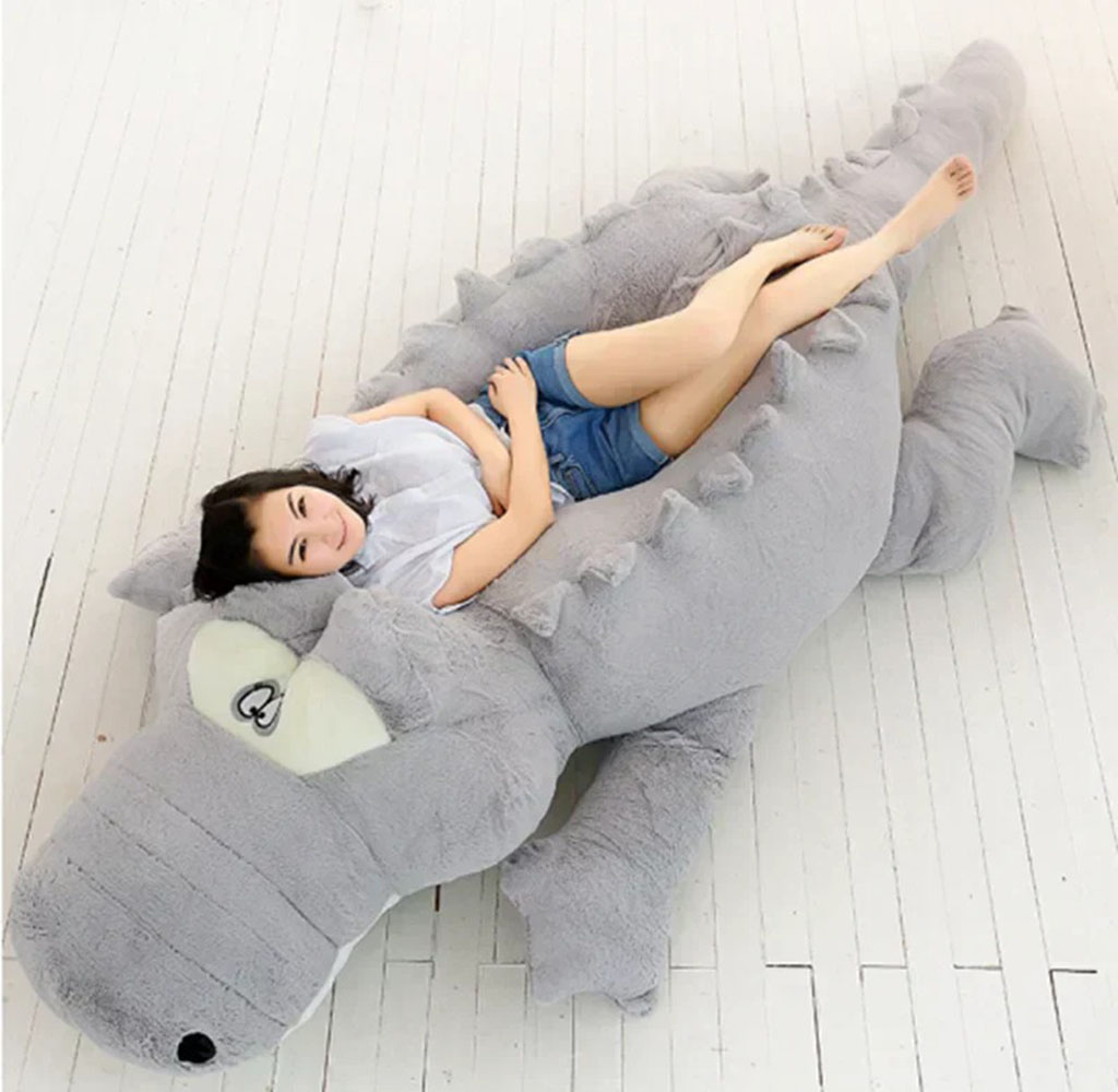 Young woman lying comfortably on an oversized gray crocodile plush toy designed as a lounger on a white wooden floor.