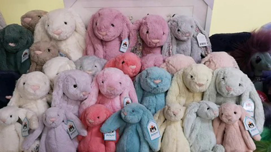 Display of soft plush bunny toys in various pastel colors, including pink, blue, cream, and gray, stacked together with tags attached.