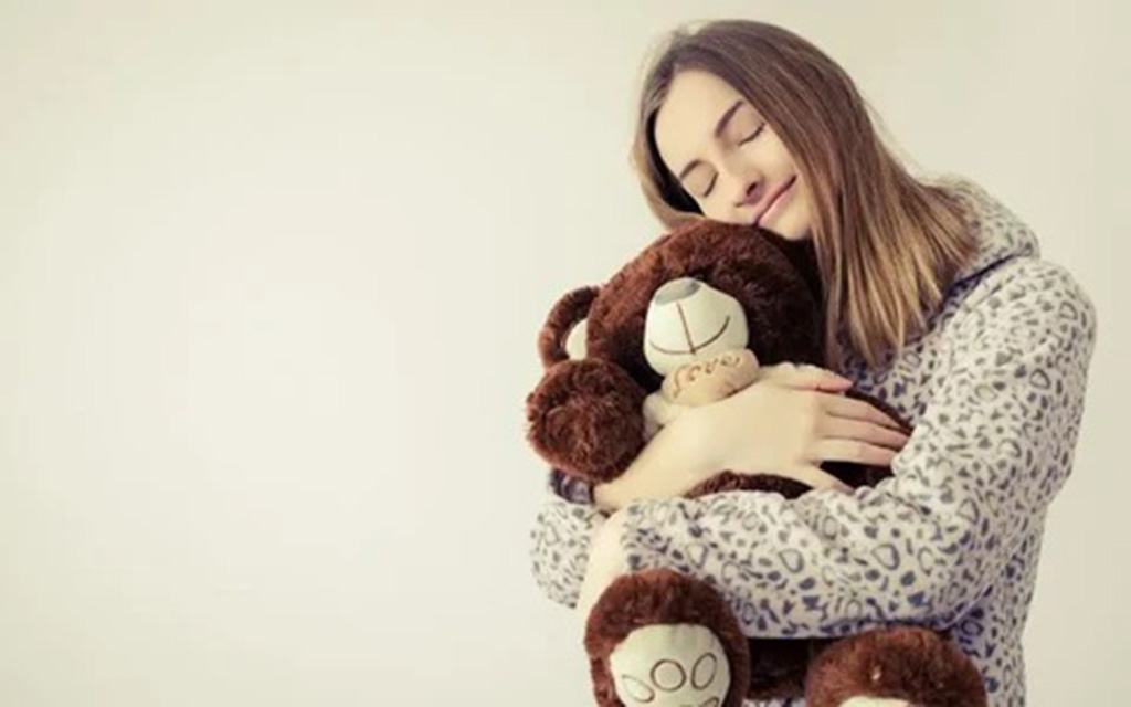 A young woman in cozy pajamas hugging a large brown teddy bear with a peaceful smile, showing the comfort and emotional support plush toys provide.