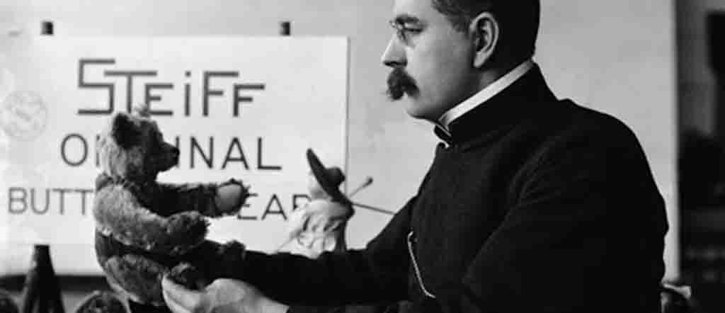 Black-and-white photo of Richard Steiff holding an early teddy bear prototype with a "Steiff Original" sign in the background.