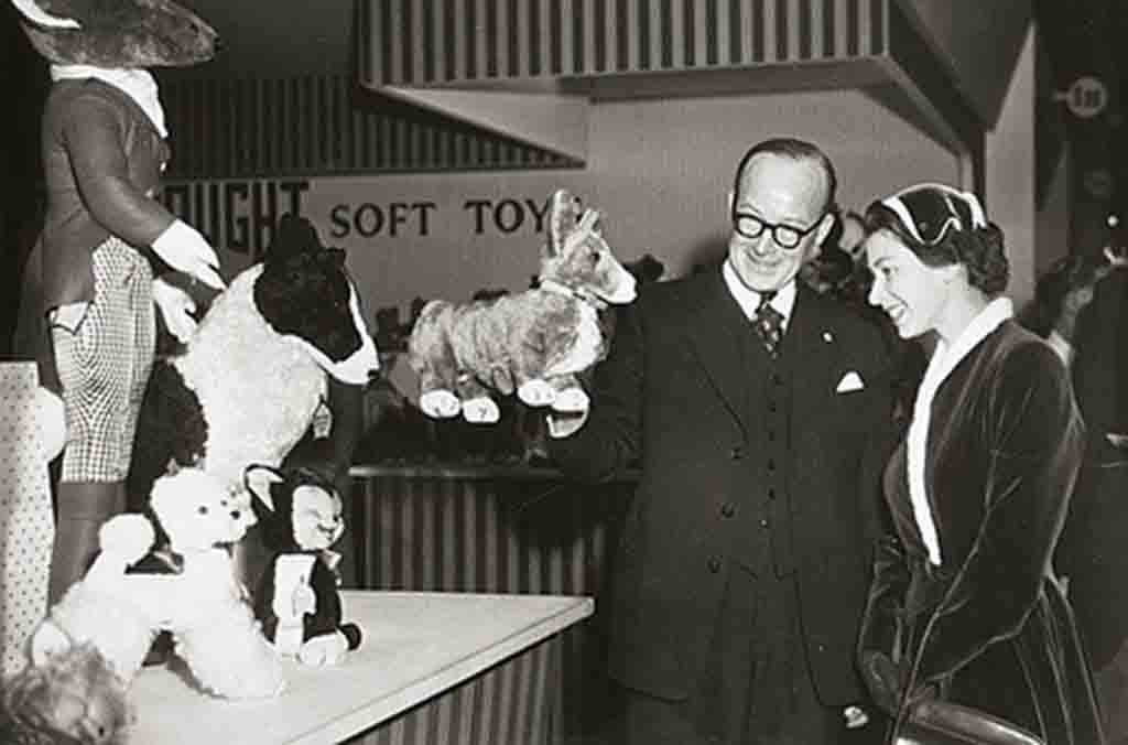 Vintage black-and-white photo showing a man and woman admiring plush animals at a Merrythought soft toy display, with teddy bears and stuffed dogs on the table.