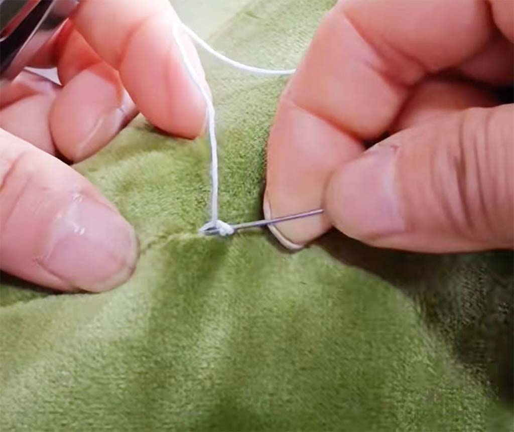 Close-up of hands stitching a green plush fabric with a needle and white thread to repair a seam.