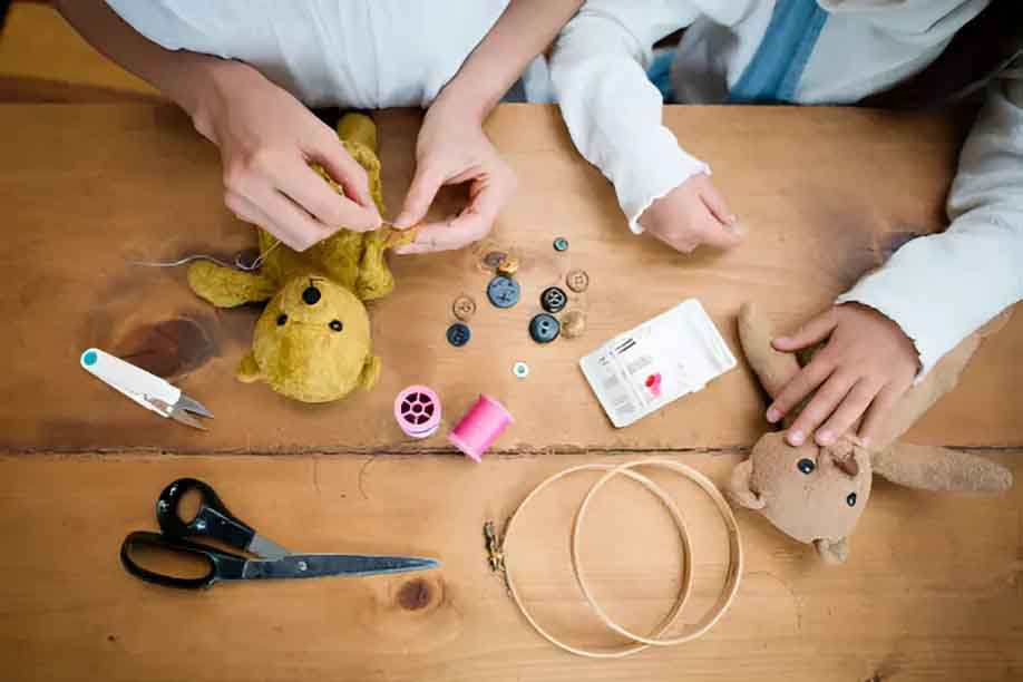 Close-up of hands sewing and repairing stuffed teddy bears on a wooden table with buttons, thread, scissors, and sewing tools.