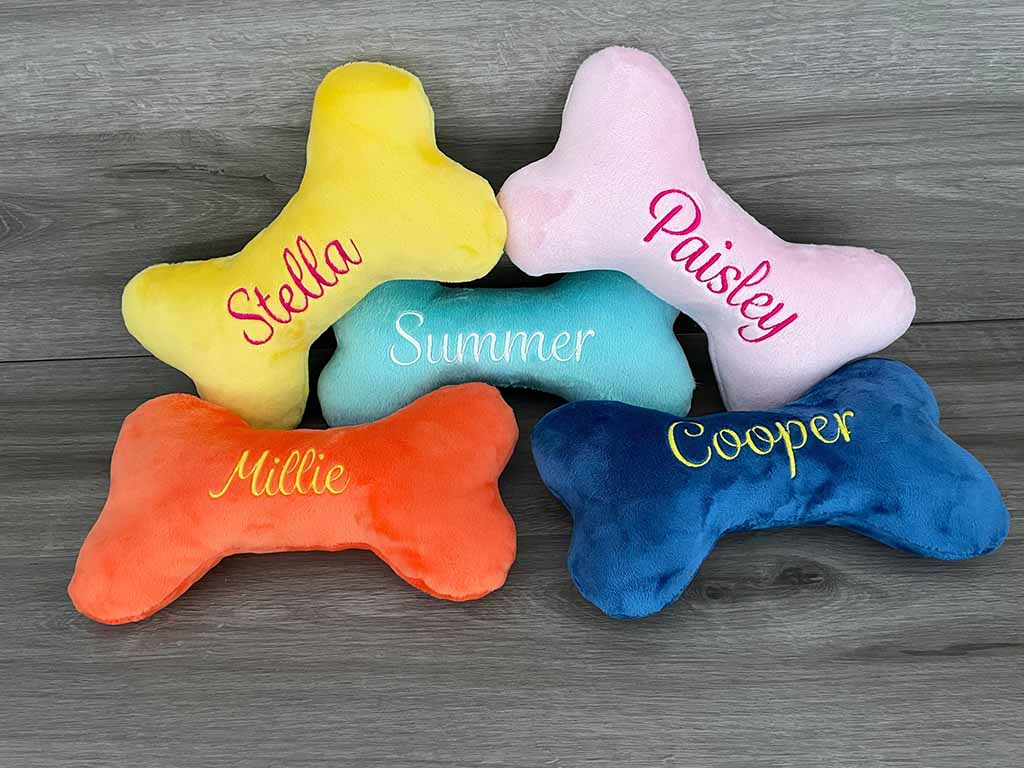 Colorful bone-shaped plush dog toys embroidered with names like Stella, Paisley, Summer, Millie, and Cooper, displayed on a wooden floor.