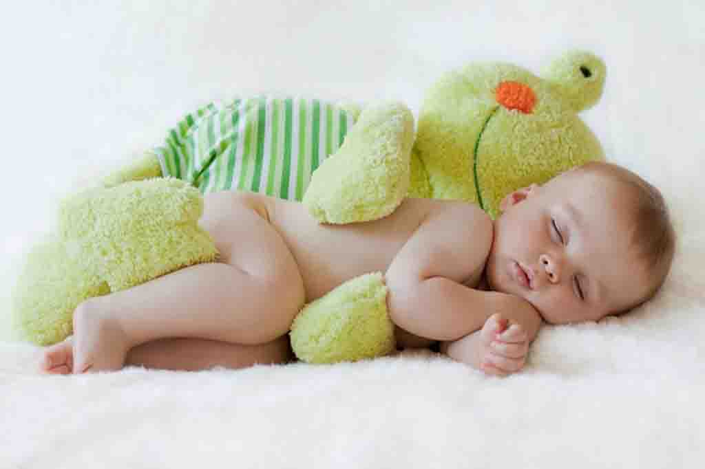 Infant lying on a soft white blanket, cuddling a large green stuffed animal while sleeping peacefully.